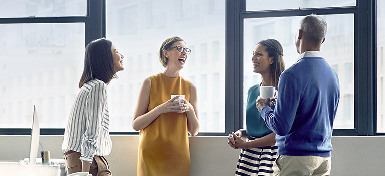 Cheerful co-workers standing by an office window, talking and laughing during a coffee break