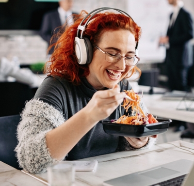 Red-headed woman with glasses eating salad at her desk while listening to music