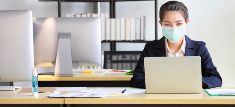 Woman wearing COVID-19 mask working in home office with hand sanitizer on desk