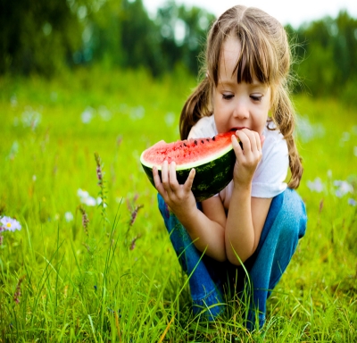 Young girl sitting in a field while eating a wedge of watermelon 