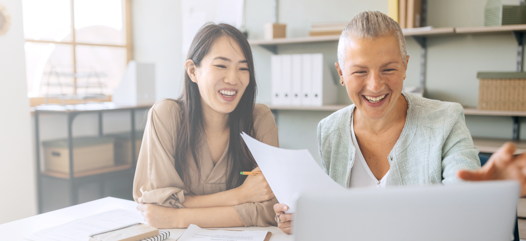 Two women holding a piece of paper and smiling while looking at a laptop.
