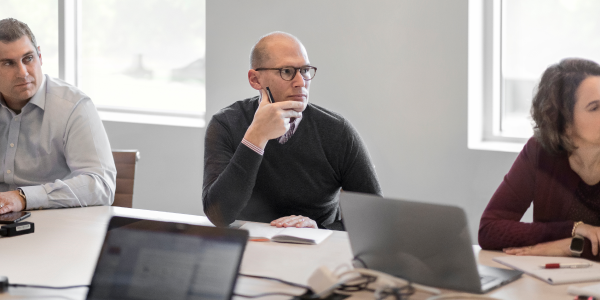 Man in a blue blazer holding a pen, engaging in a discussion with a colleague, with documents and a laptop on the table.