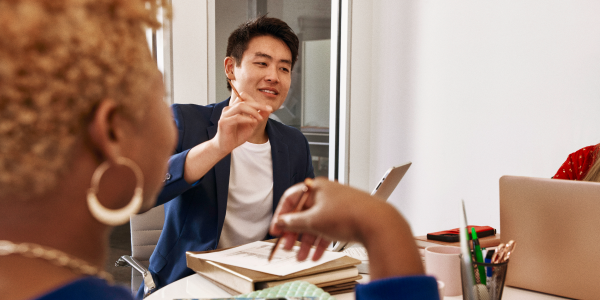 Three colleagues in a conference room. Two men are shaking hands and smiling, while a woman stands nearby holding a notebook and smiling. A laptop and documents are on the table.
