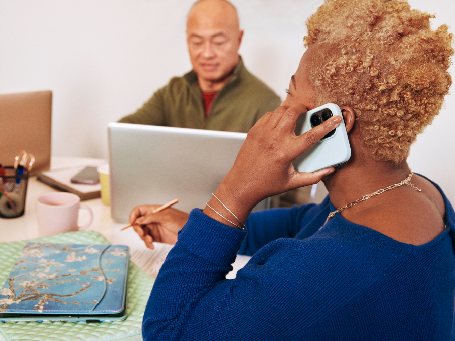 Two colleagues in an office. One is on a smartphone, the other on a laptop.