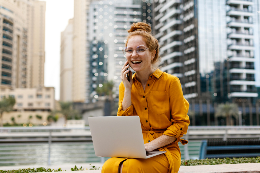 Woman on phone and laptop with building in the background