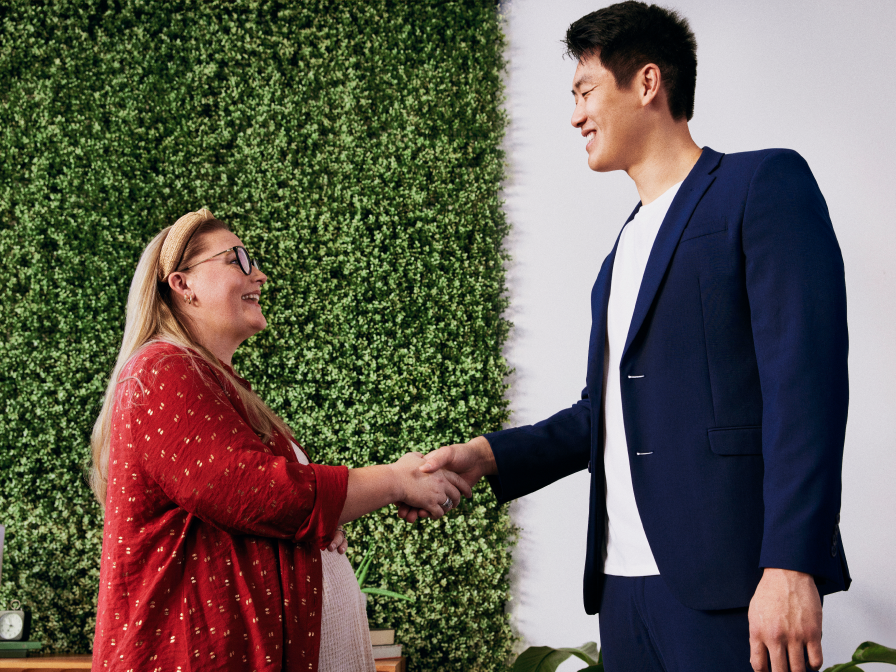 A woman with long blonde hair, wearing glasses and a red cardigan, smiling and shaking hands with a man in a navy blue suit.